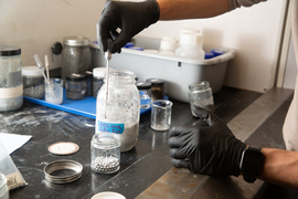 Gloved hands mix a slurry of ingredients in a mason jar in the lab.